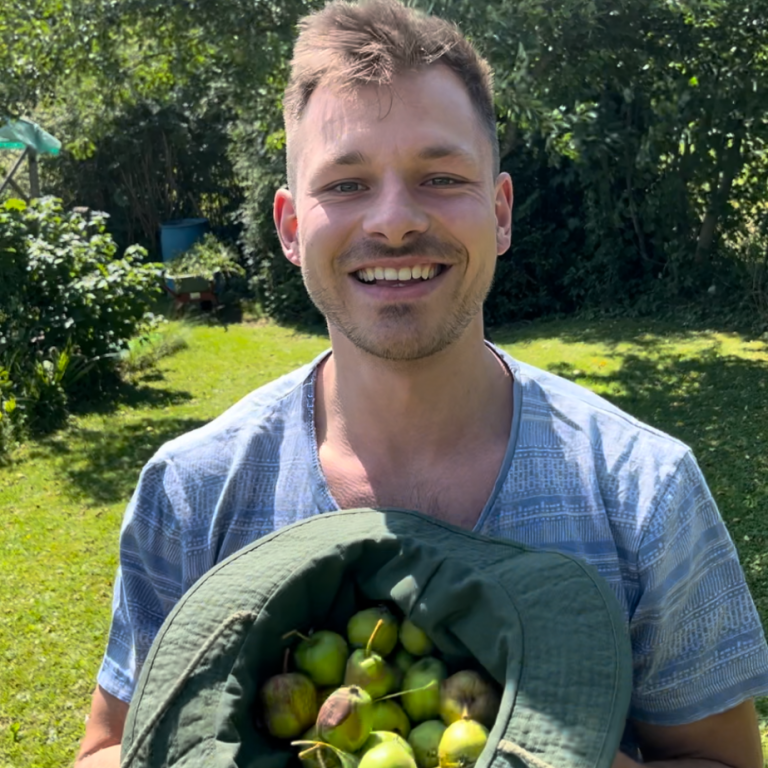 Junger Mann im Freien, der mit einem Hut voll Äpfeln lächelt.
