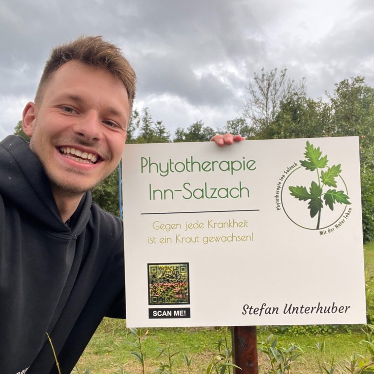 Lächelnder Mann mit Pflanzenheilkunde Schild Lächelnder Mann steht neben einem Schild für die Physiotherapie Inn-Salzach.